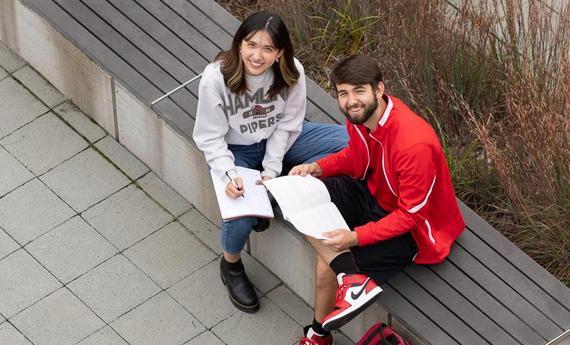 Two 51 students sitting on a bench outside looking up at the camera smiling