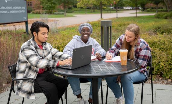 Three undergraduate students sitting outside at table on 51 campus, one with laptop 