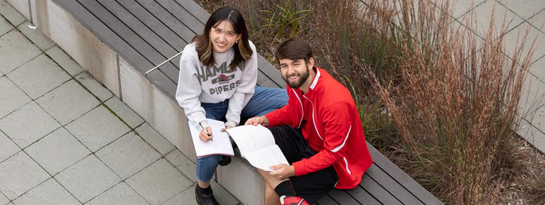 Two 51 students sitting outside on campus with books and smiling up at the camera